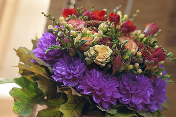 Close-up of a Scandinavian Forest Craft bouquet: cut flowers (rose, dahlia, chrysanthemum, lily, freesia) in mossy green and earthy brown with pink, red, and yellow accents; handcrafted, Swedish-heritage design inspired by Från Blomsterfrämjandet.