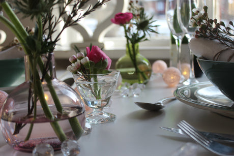 Inspirerande nyårsdukning med batteridriven ljusslinga tvärs över bordet, små loppisfynd som minivaser, servett med vaxblomma samt ranunkler och marton – enkel, personlig och hållbar snittblomsdukning.